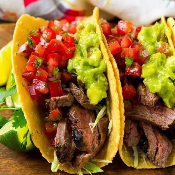 Two steak tacos with guacamole, pico de gallo, and shredded lettuce on a wooden board.