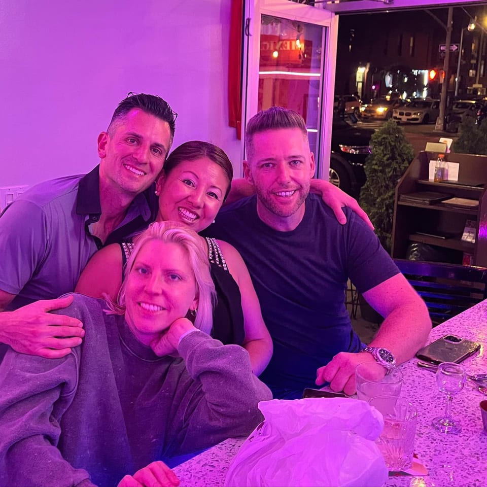 Four smiling friends pose together at a bar counter with purple lighting at Fusion Bar and Grill.