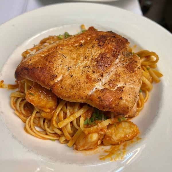 Grilled fish fillet over linguine pasta with a red sauce and seafood, garnished with herbs.
