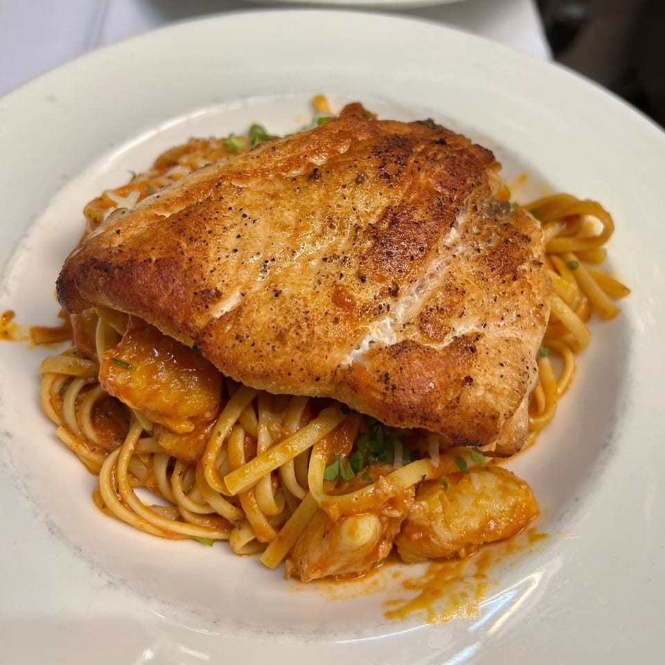 Grilled fish fillet over linguine pasta with a red sauce and seafood, garnished with herbs.