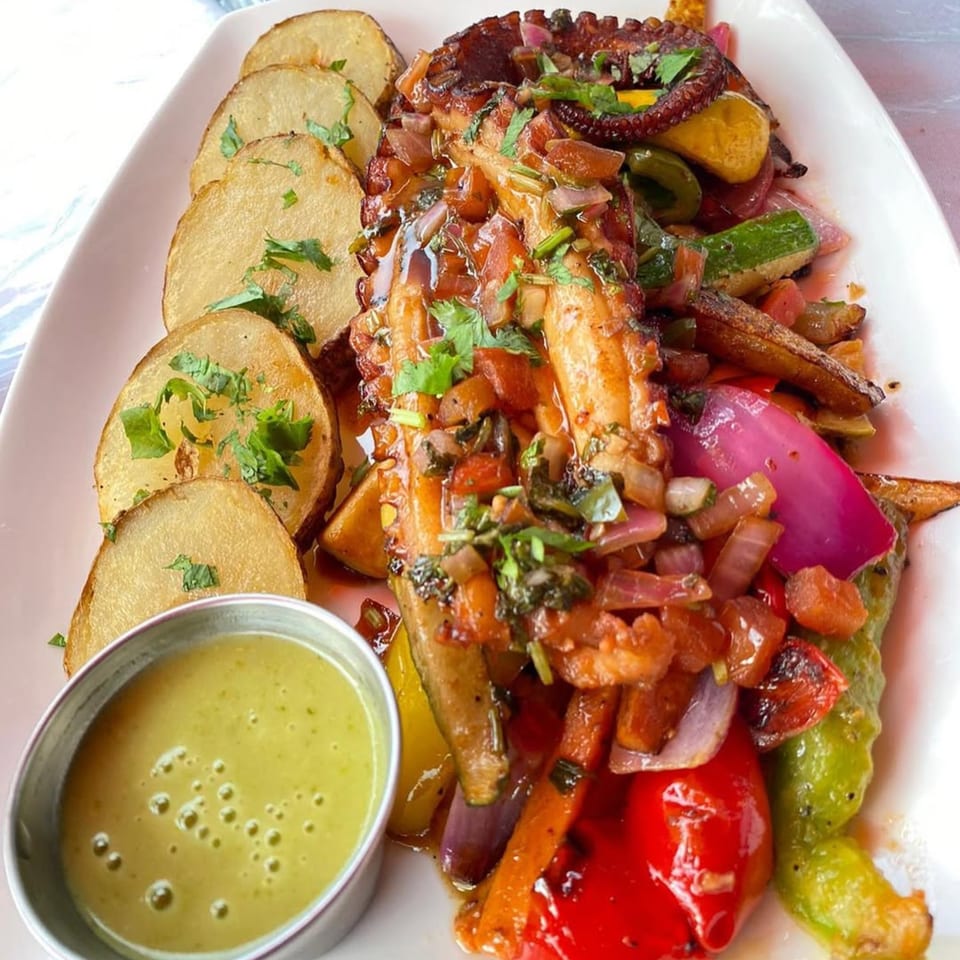 Grilled octopus with roasted vegetables, sliced potatoes, and a side of green sauce on a white plate.